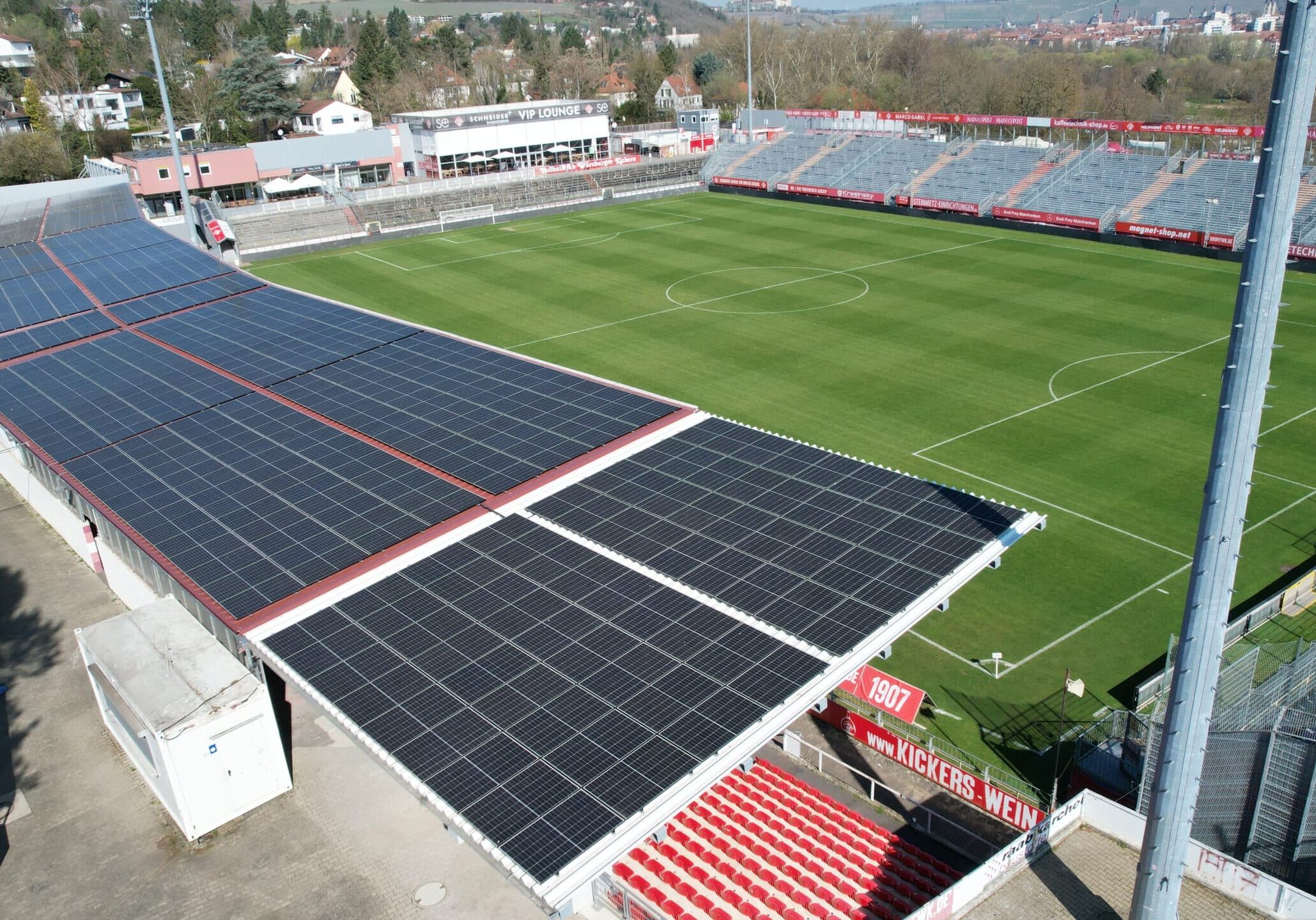 Fußballstadion mit grüner Rasenfläche und leeren Tribünen, auf den überdachten Zuschauerbereichen großflächig installierte Photovoltaikanlagen.