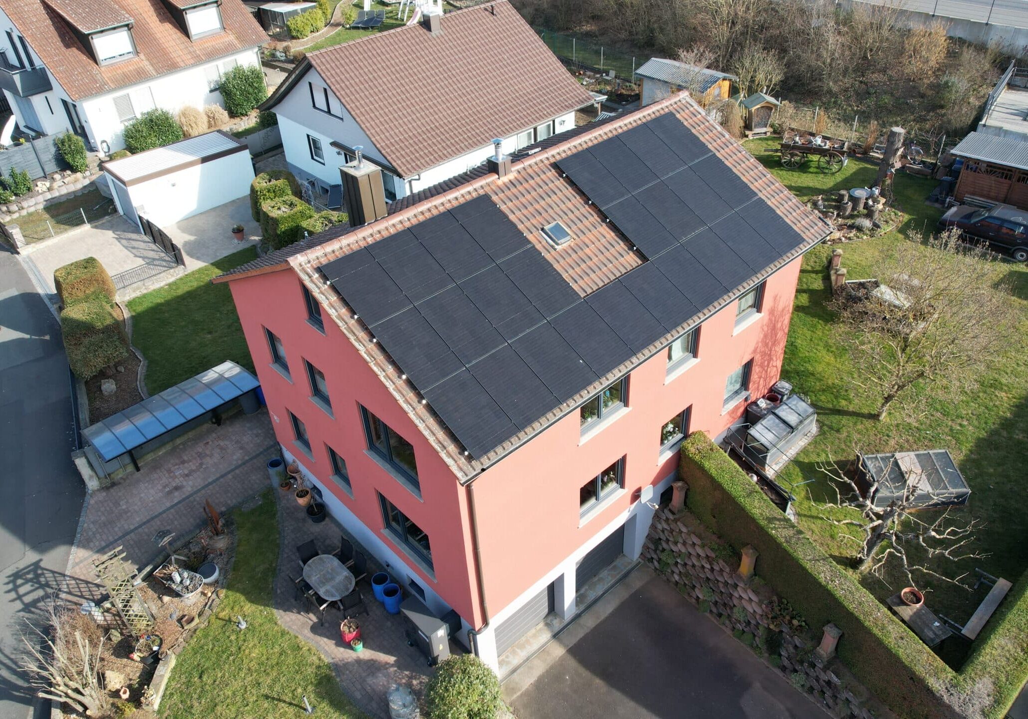 Mehrfamilienhaus mit roter Fassade und dunklem Satteldach, ausgestattet mit Photovoltaikanlage, Blick auf umliegende Grundstücke und Gärten.