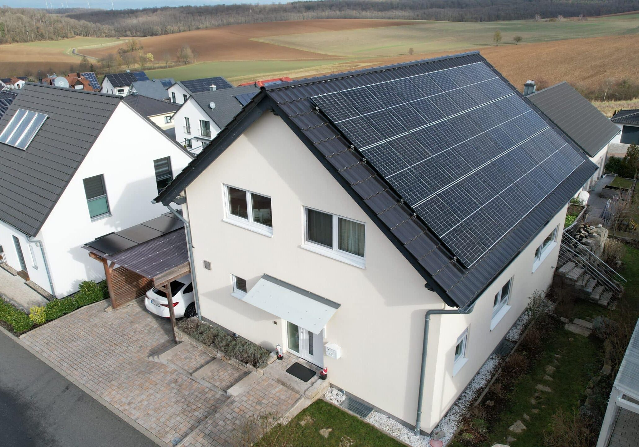 Einfamilienhaus mit dunklem Satteldach und installierter Photovoltaikanlage, Blick auf gepflasterte Einfahrt, Garten und angrenzende WohnhÃ¤user.
